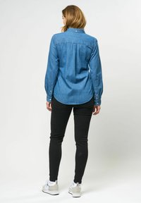 Denim overhemd met lange mouwen, lichtblauwe kleur, knoopdetails en een gebogen zoom, gecombineerd met zwarte broek en witte sneakers.