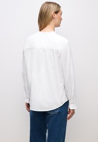 Hvid langærmet bluse med en flæseudskæring og rynket rygdetalje, parret med blå denimjeans med en afslappet pasform.