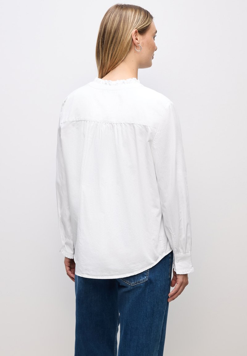 Hvid langærmet bluse med en flæseudskæring og rynket rygdetalje, parret med blå denimjeans med en afslappet pasform.