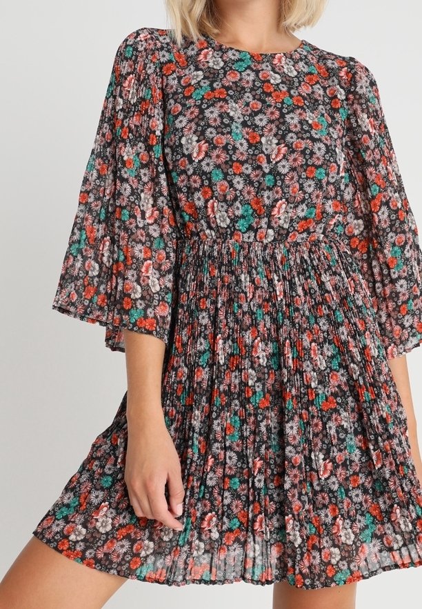 Femme portant une robe plissée à fleurs rouges, vertes et blanches sur fond noir, avec des manches trois-quarts amples.