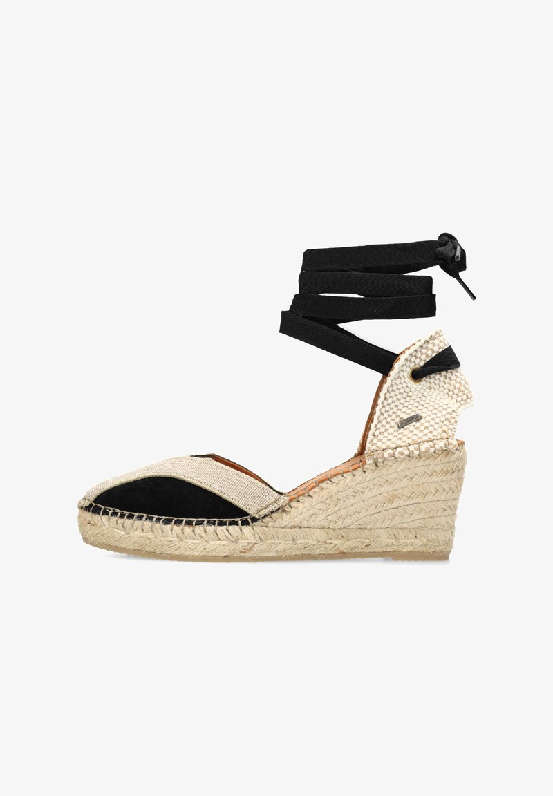 La sandaletta con zeppa espadrille presenta una suola in juta intrecciata, accenti in tessuto nero, tomaia in tela beige e lacci neri che si incrocciano alla caviglia.