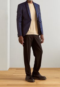 Blazer a fantasia navale, maglione beige e pantaloni neri. Il blazer presenta un design elaborato, con tessuto lucido e vestibilità su misura. Scarpe nere con suola spessa.