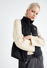 Gilet rembourré noir avec un col montant, fermeture éclair et patch logo, associé à un pull en crème tricoté présentant une texture à mailles torsadées.