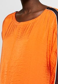 Orangefarbene, strukturierte Bluse mit einem Bootsausschnitt und Falten. Mit schwarzen Akzenten an der Schulter und weißen Paspeln an der Seite.