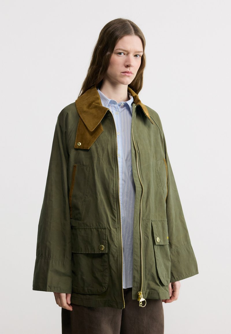 Νεαρή γυναίκα με μακριά, λυτά μαλλιά φοράει μια φαρδιά ελιά πράσινη ζακέτα με καφέ κολάρο από κορντόρι και μεγάλες μπροστινές τσέπες, στέκεται μπροστά σε λευκό φόντο.