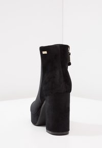 Bottine en daim noir avec talon haut effilé, dotée d'une fermeture éclair latérale et d'un petit accent métallique près du talon. Texture lisse, design minimaliste.