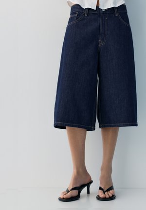 Oseba, oblečena v temno modre jeans culottes hlače in črne natikače s peto, stoji ob svetlo sivi ozadju.