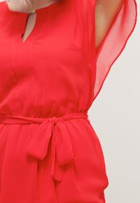 Vestido rojo ligero con escote en forma de ojo de cerradura y mangas cortas y fluidas. Presenta un cinturón con lazo que acentúa la forma y el estilo.