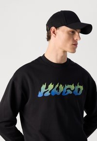 Young man wearing a black cap and black sweatshirt with blue and green flame-style "HUGO" text on the chest, facing sideways.