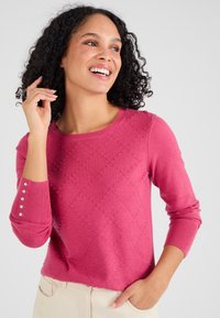 Roze gebreide trui met een ruitpatroon, geribbelde manchetten en decoratieve knopen op de mouwen. Glad textuur en ronde ronde hals.