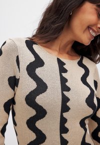 Kootud sviiter, millel on beež baaspind mustade sakilise mustritega, ümar krae ja pikad varrukad. Pehme tekstuur ja lihvitud disain.