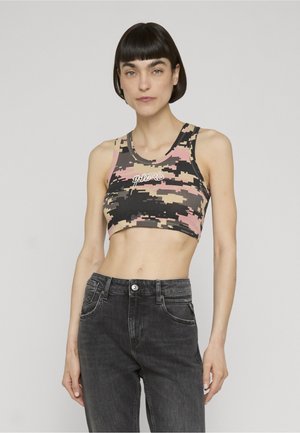 Vrouw met kort donker haar, draag een meerkleurige digitale camouflagetop en een zwarte hoge taille spijkerbroek tegen een effen achtergrond.