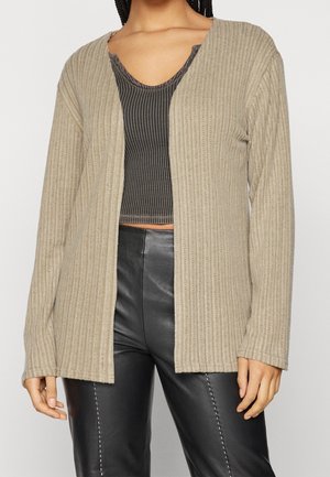 Žena nosící béžový žebrovaný kardigan s otevřeným předním dílem, černý crop top s vzorem a černé kožené kalhoty, stojící před jednoduchým pozadím.