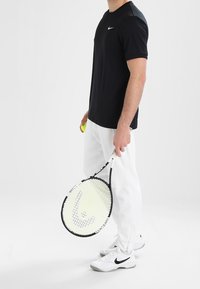 Camiseta atlética negra de Nike, pantalones blancos, zapatos blancos de Nike, sosteniendo una raqueta de tenis con un mango amarillo y negro, mostrando un diseño de carita sonriente.