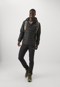 Veste noire matelassée avec une fermeture éclair, associée à un pantalon noir et des bottes de randonnée brunes. Elle possède une capuche intégrée et un léger sac à dos.