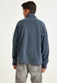 Fleecetröja i mörkblått med hög krage, avslappnad passform, långa ärmar och texturerad tyg. Buren med grå denimbyxor, bakvy visas.