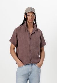Kortærmet, knappet skjorte i et struktureret mauve stof. Sættes sammen med lyseblå denimjeans og en beige kasket med et simpelt logo.