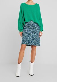 Suéter verde de punto con hombros caídos, combinado con una falda azul de estampado floral y botas estilo occidental blancas hasta el tobillo.