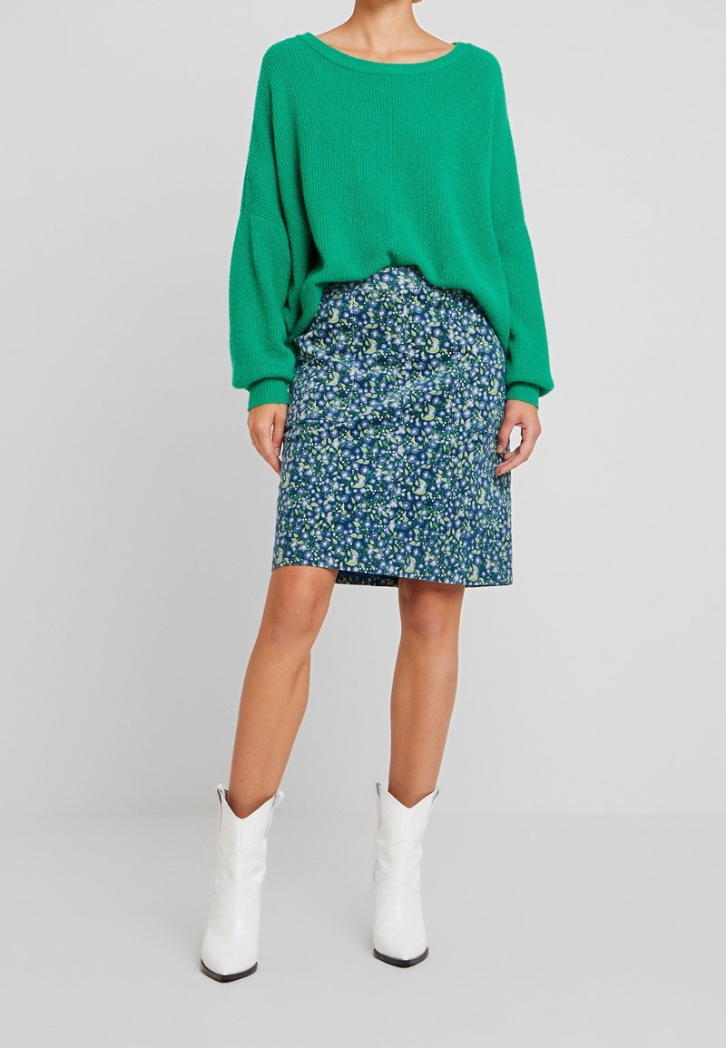 Suéter verde de punto con hombros caídos, combinado con una falda azul de estampado floral y botas estilo occidental blancas hasta el tobillo.