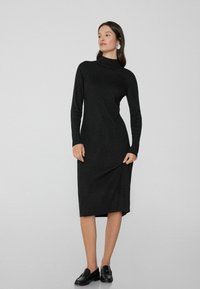 Schwarzes, langärmliges, Turtleneck-Kleid aus geripptem Stoff, knielang mit lockerer Passform. Kombiniert mit schwarzen Schuhen und minimalem Schmuck.