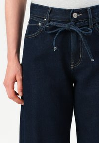 Tmavě modré džíny s elastickým pasem na šňůrku. Mezi vlastnosti patří výrazné prošití, pět kapes a mírně uvolněný střih.