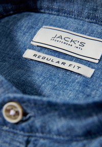 Collo della camicia in denim con una texture azzurro chiaro; le etichette riportano "JACK'S SPORTSWEAR INTL" e "REGULAR FIT", con un dettaglio di un bottone.