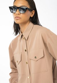Camicia beige con bottoni, due tasche sul petto, accessori in oro e un tessuto liscio e leggero; abbinata a occhiali da sole neri angolari.