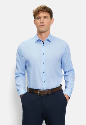 Mann trägt ein hellblaues Hemd, das in dunkelblaue Hosen mit braunem Ledergürtel gesteckt ist, steht mit den Händen in den Taschen.