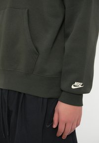 Alyvuogių spalvos džemperis su priekinėmis kišenėmis ir galuotos rankovėmis, kairėje rankovėje turi baltą išsiuvinėtą Nike logotipą. Juodi sportiniai šortai su mažuoju juodu Nike logotipu.