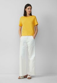 T-shirt en coton jaune à manches courtes, associé à un pantalon large blanc. Le modèle se tient les mains dans les poches, portant des sandales blanches.