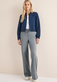 Femme blonde portant une veste zippée bleu marine, un haut blanc, un pantalon à cordon gris et des baskets blanches, se tenant devant un mur beige.