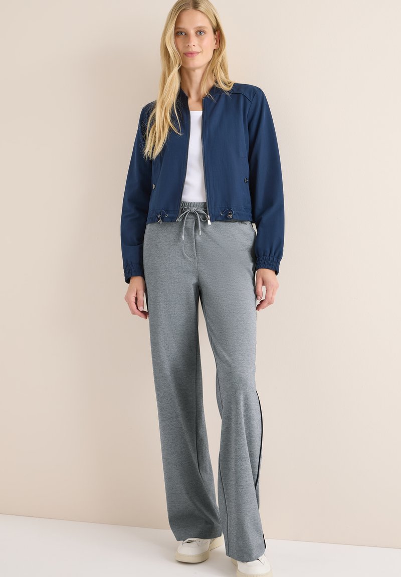 Femme blonde portant une veste zippée bleu marine, un haut blanc, un pantalon à cordon gris et des baskets blanches, se tenant devant un mur beige.