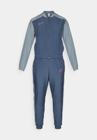 Nike træningsdragt med blålig krop, grå ærmer, lynlås foran, elastisk talje og et rødt logo på brystet og hoften. Polyester materiale.
