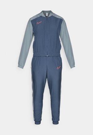 Nike treningsdrakt med en marineblå kropp, grå ermer, glidelås foran, elastisk midjebånd, og en rød logo på brystet og hoften. Polyester materiale.