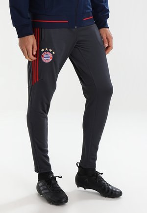 Mężczyzna ubrany w ciemnoszare spodnie piłkarskie FC Bayern München z czerwonymi paskami i czarne korki, stojący na białej podłodze.