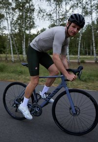 Grijze racefiets met een lichtgewicht frame, zwarte wielen en schijfremmen. Fietser in grijze trui en donkergroene shorts, witte sokken en schoenen.