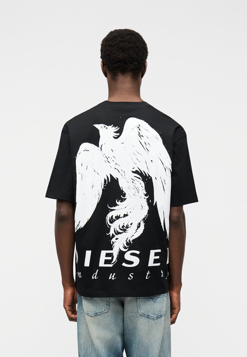 Άτομο που φοράει μαύρο T-shirt με μεγάλο λευκό γραφικό φοίνικα και το κείμενο «DIESEL industry» στην πλάτη, συνδυασμένο με ανοιχτόχρωμο μπλε τζιν.