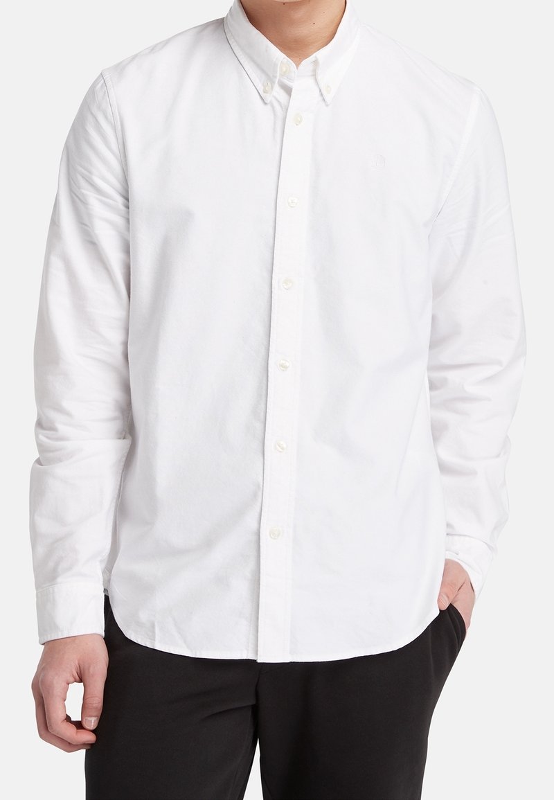 Homme portant une chemise blanche unie à manches longues boutonnée avec un pantalon noir, debout, une main dans la poche.