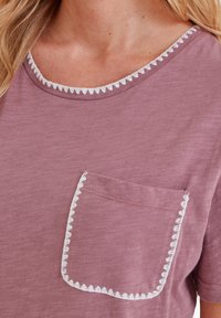 Close-up of mauve t-shirt with white scalloped trim on neckline and pocket, worn by person with long blonde hair.