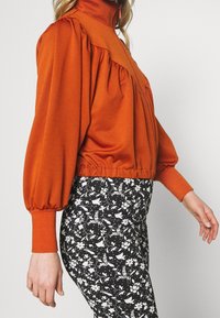 Sudadera naranja con cuello alto, hombros fruncidos y dobladillo elástico, combinada con leggings negros con estampado floral.