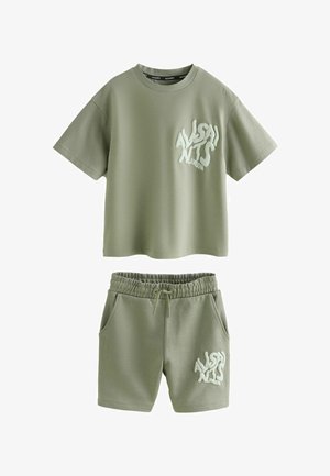 Svijetlozeleni set od pamučne majice i kratkih hlača s grafičkim printom, opuštenog kroja i okruglim izrezom. Nošen s bijelim tenisicama i čarapama.