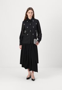 Camicia nera a maniche lunghe con piccole decorazioni argento, abbinata a una gonna nera plissettata, tiene una pochette grigia testurizzata e indossa tacchi neri.