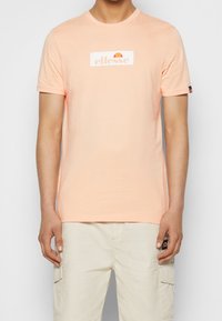 Krátký rukáv, tričko s kulatým výstřihem z měkkého broskvového materiálu, se středovým bílým logem a oranžovou grafikou. Nosené s světle béžovými cargo šortkami.