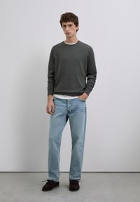 Grauer Pullover, blaue Jeans, braune Schuhe. Der Pullover hat einen Rundhalsausschnitt und lange Ärmel; die Jeans sind locker sitzend mit einer ausgewaschenen Optik.