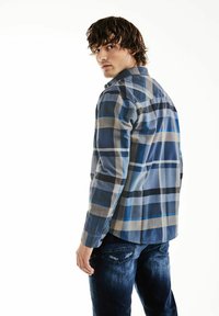 Camicia a quadri con motivi blu, grigi e neri, maniche lunghe e dettagli con bottoni. Indossata con jeans scuri, vista da dietro.