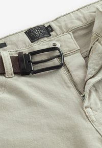 Pantalones beiges con cremallera parcialmente abierta, cinturón negro con hebilla de metal y etiqueta que muestra la marca "Next" y la talla 32R.