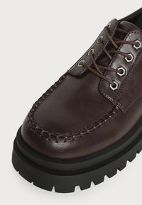 Chaussure en cuir marron foncé avec une semelle épaisse noire à crampons, couture mocassin et lacets marron passés dans des œillets métalliques.
