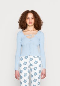 Top de manga longa em azul claro com nervuras, decote em V e detalhe de laço. Combinado com calças brancas com grandes padrões de flores azuis.