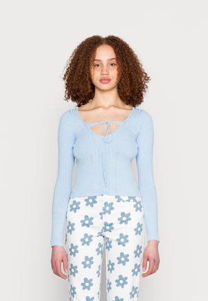 Top de manga longa em azul claro com nervuras, decote em V e detalhe de laço. Combinado com calças brancas com grandes padrões de flores azuis.