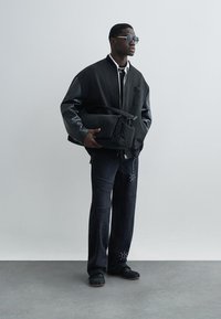 Fekete bombázó dzseki bőrszerű ujjakkal, fehér ing, kopott fekete farmer gyöngy díszítéssel és fekete sportcipő, kezében fekete táskával.